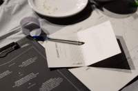 Table with three different printed materials, a pen, a white paper strip with zigzag edges and the edge of a plate. The printed materials are either white or black, and have text printed on.