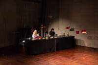 Two people sit behind a long table, with several objects. The person on the right has their laptop open on the table. Two structures hang from the ceiling to the right of the table.