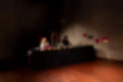 Two people sit behind a long table, with several objects. The person on the right has their laptop open on the table. Two structures hang from the ceiling to the right of the table.