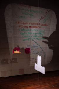 A transparency sheet with handwritten notes is being projected on a tiled wall. The larger sentence, in red, reads: 