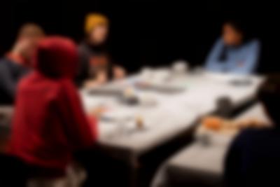 Five people sit around two rectangular tables, engaging in conversation. There is a spotlight shining on the table; the space around it is in the dark. Bowls, plates, mugs and cutlery are set on the table. A board with bread and dip is partially visible.