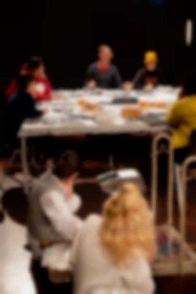 Five people sit around a table. The person facing the camera gesticulates with their hands, while the others look in their direction. There is food on the table, as well as cutlery, crockery and other paper materials. Two people sit on the floor, also looking towards the person gesticulating.