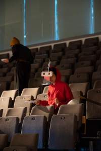 A person sits in tribute seating, wearing a headset and holding a plate. Another person walks behind them.