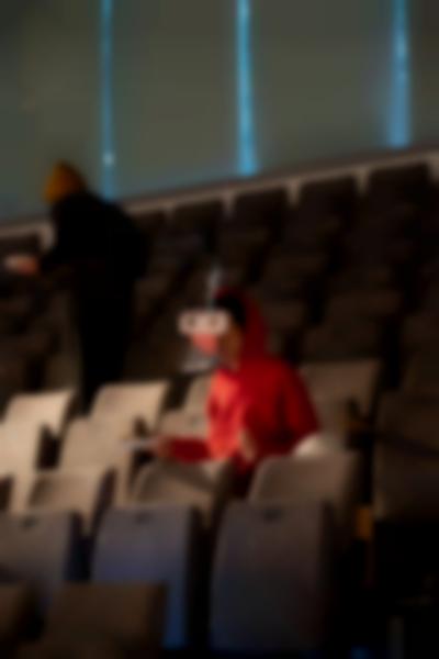 A person sits in tribute seating, wearing a headset and holding a plate. Another person walks behind them.